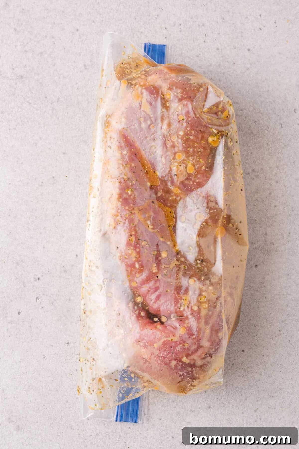Raw marinated meat sealed in a clear plastic ziplock bag, placed on a light gray textured surface.