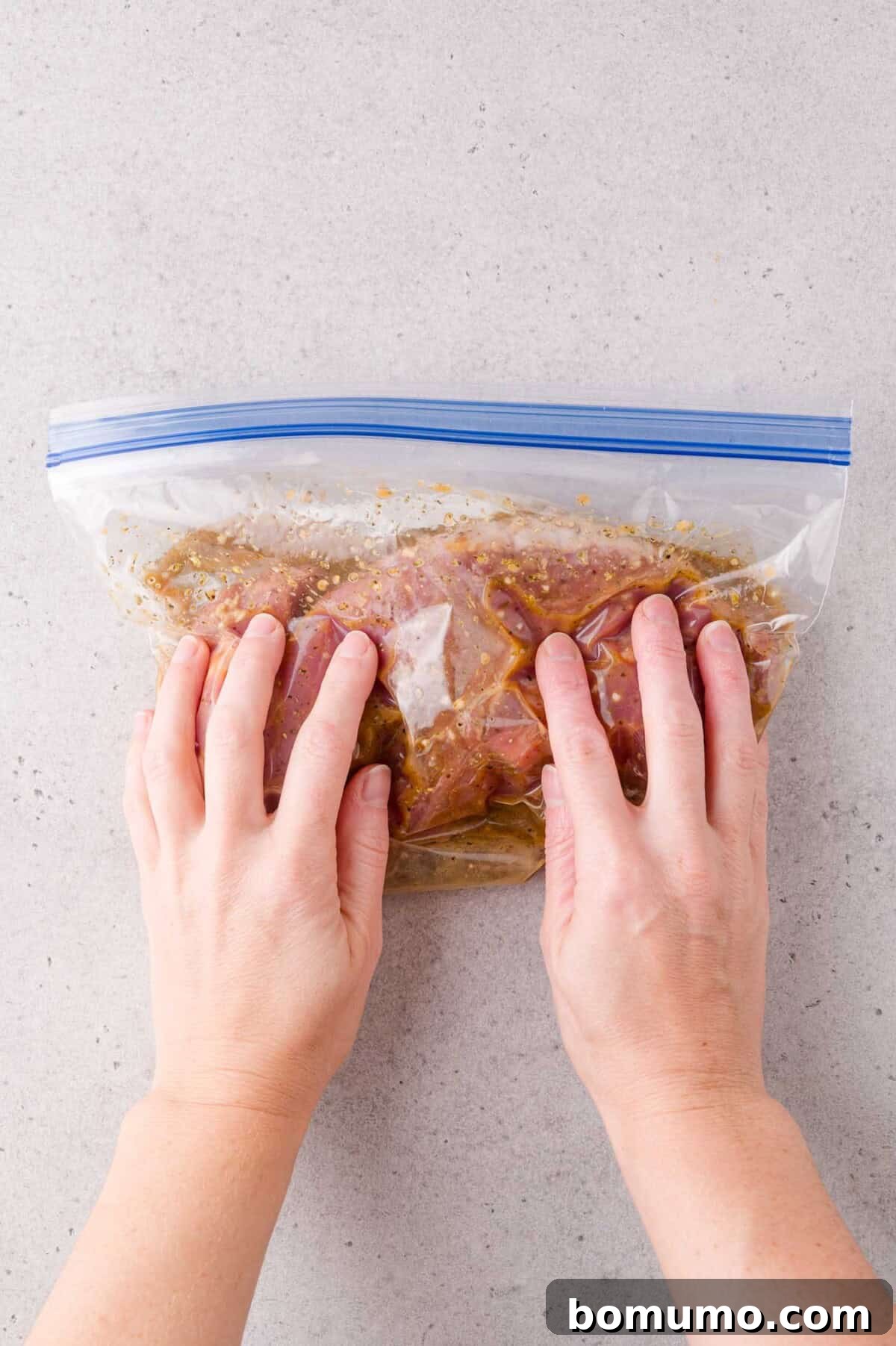 Hands sealing a plastic zip-top bag containing marinated meat on a light-colored countertop.