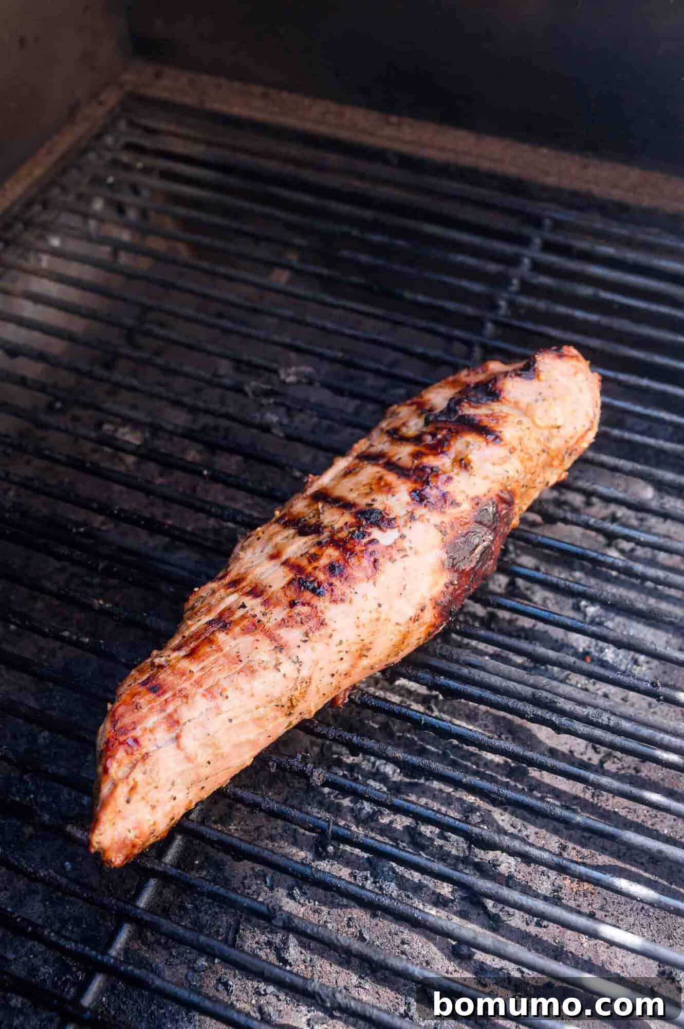 A seasoned pork tenderloin cooking on a grill, showing visible grill marks and slight charring.