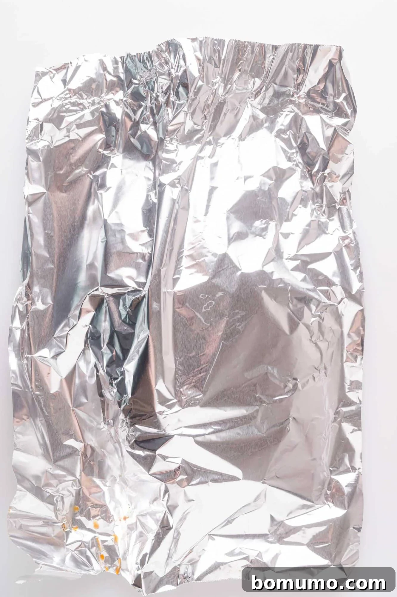 A crumpled sheet of aluminum foil with a few small orange crumbs on a white background.
