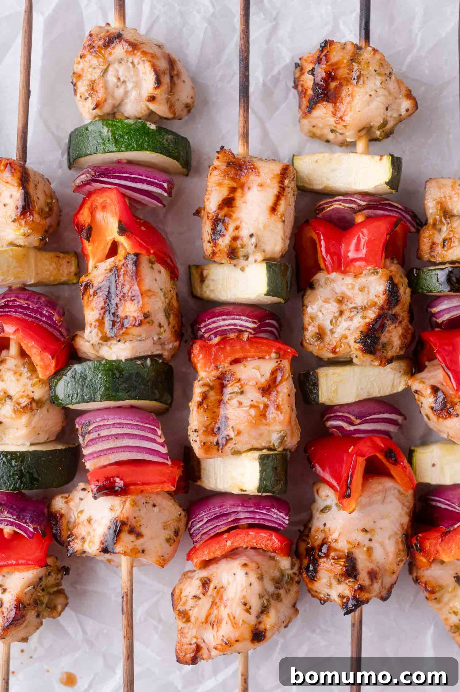 Grilled chicken kebabs with pieces of zucchini, red bell pepper, and red onion on wooden skewers, arranged on a white surface.