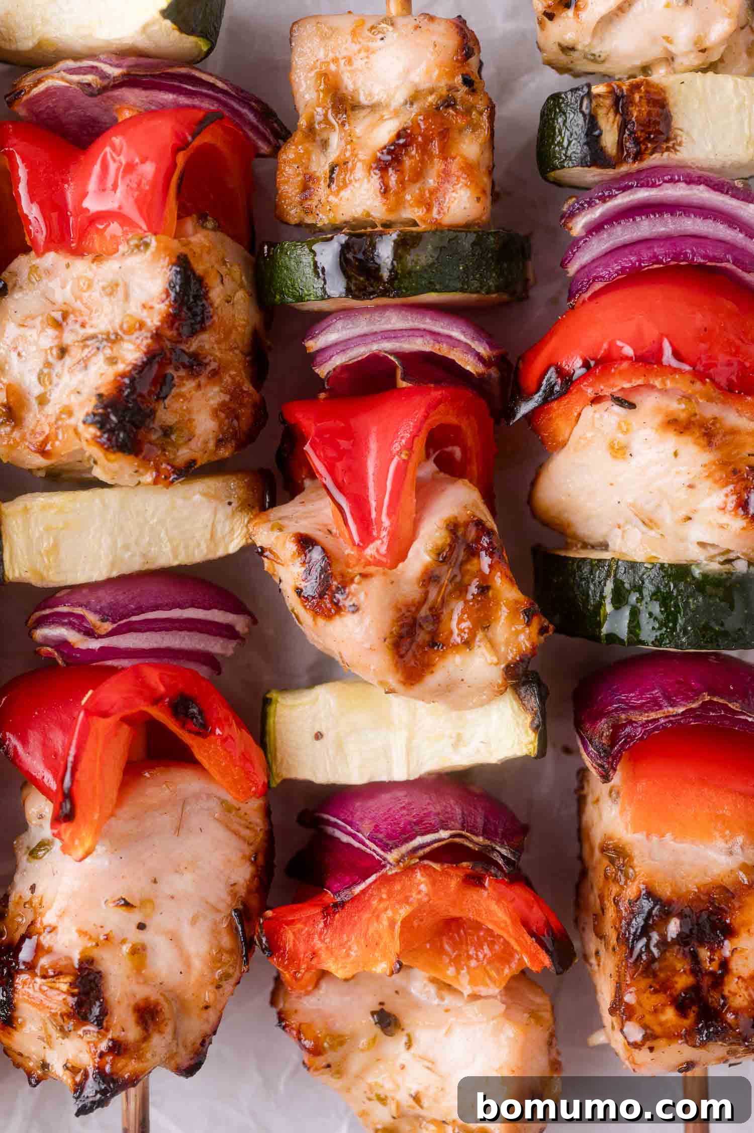 Grilled chicken skewers with pieces of red bell pepper, zucchini, and red onion on wooden sticks, arranged closely together.