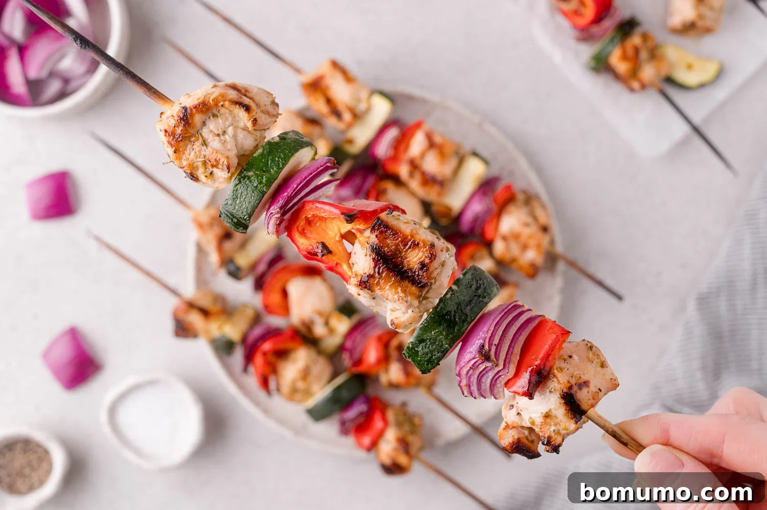 A hand holds a grilled skewer with pieces of chicken, zucchini, red onion, and red bell pepper over a plate of similar skewers on a light surface.