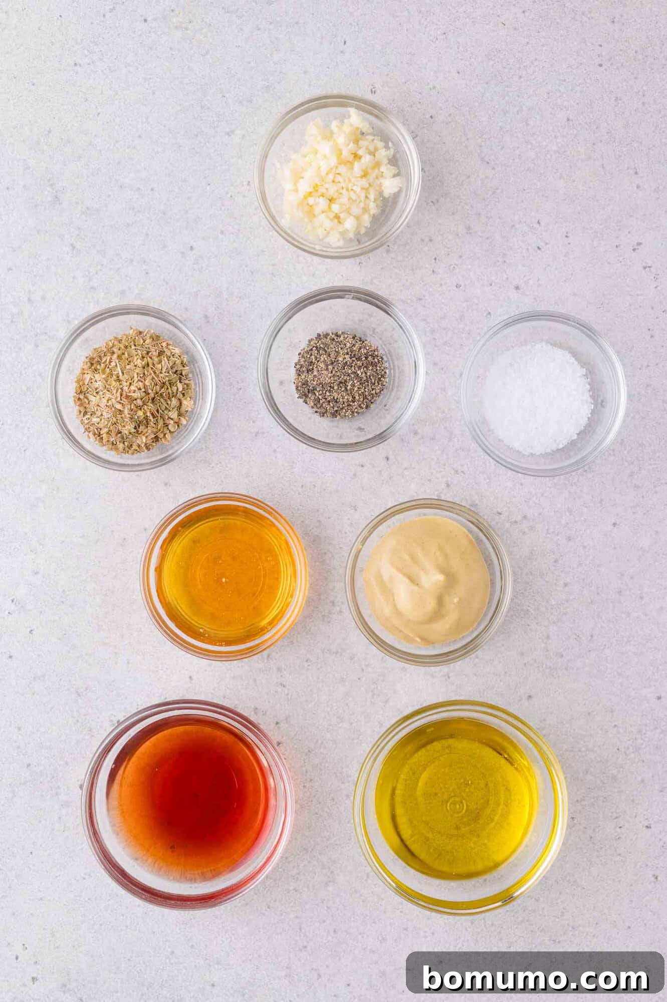 Nine small glass bowls containing minced garlic, dried herbs, black pepper, salt, honey, Dijon mustard, vinegar, and olive oil are arranged on a light surface.