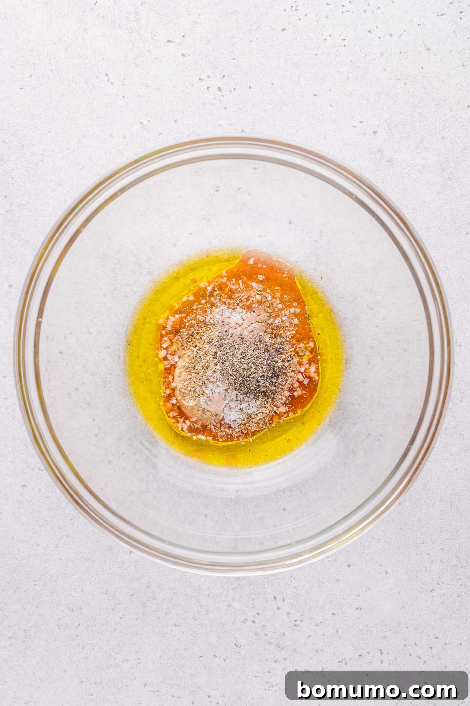 A clear glass bowl containing olive oil, honey, and seasonings on a light countertop.