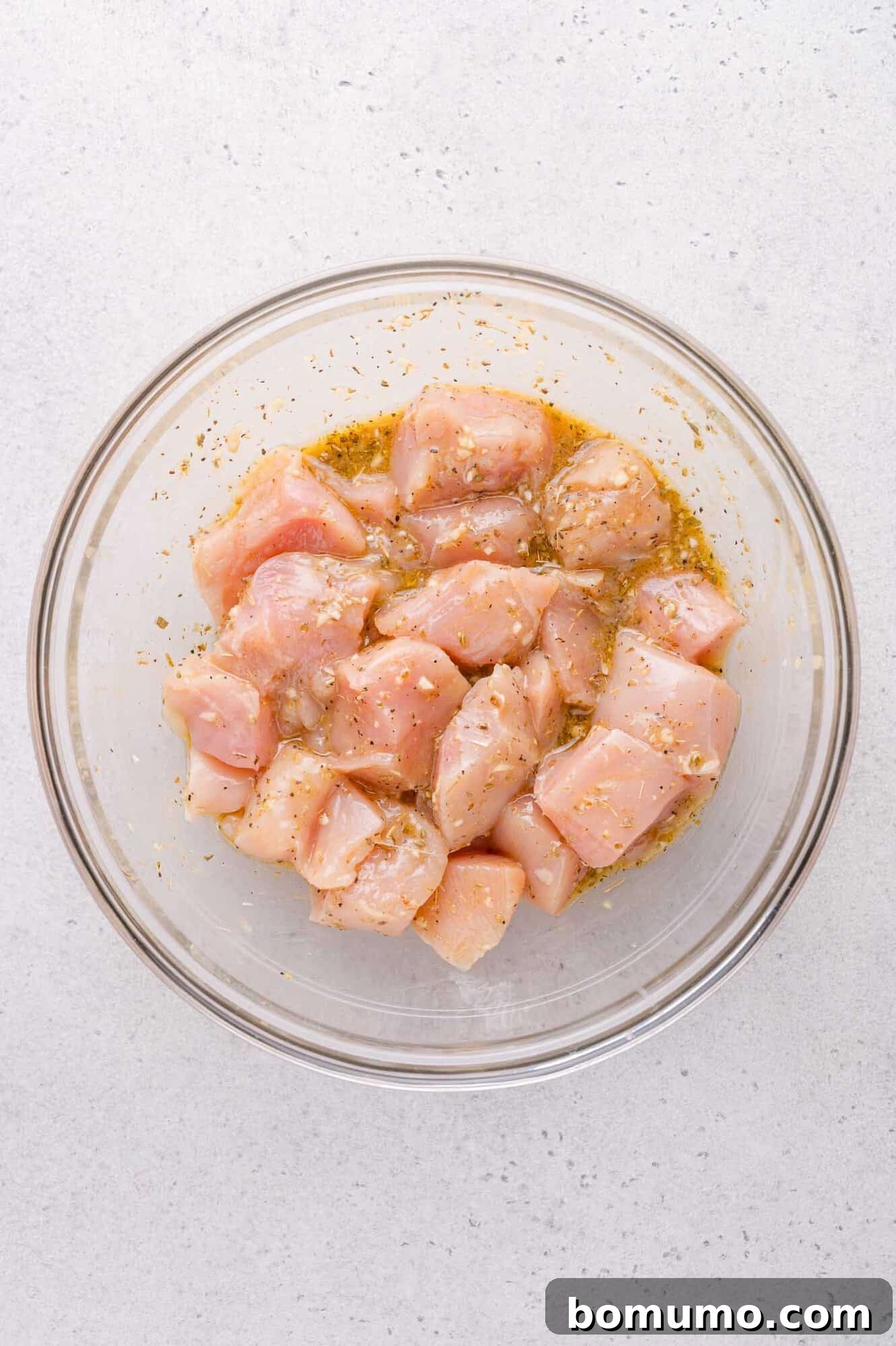 A glass bowl filled with raw, cubed chicken marinating in oil and seasonings on a light-colored surface.