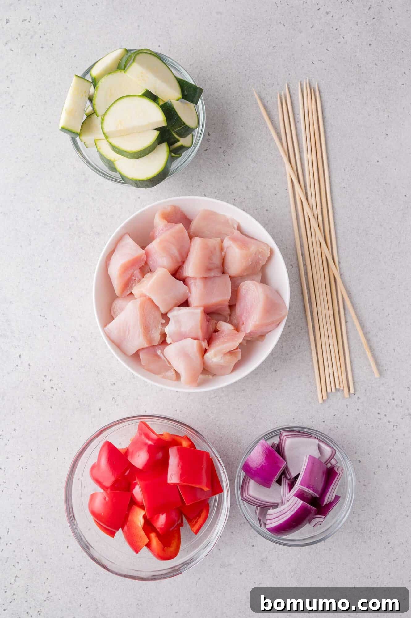 Bowls of chopped zucchini, raw chicken, red bell pepper, and red onion with wooden skewers arranged on a light surface.