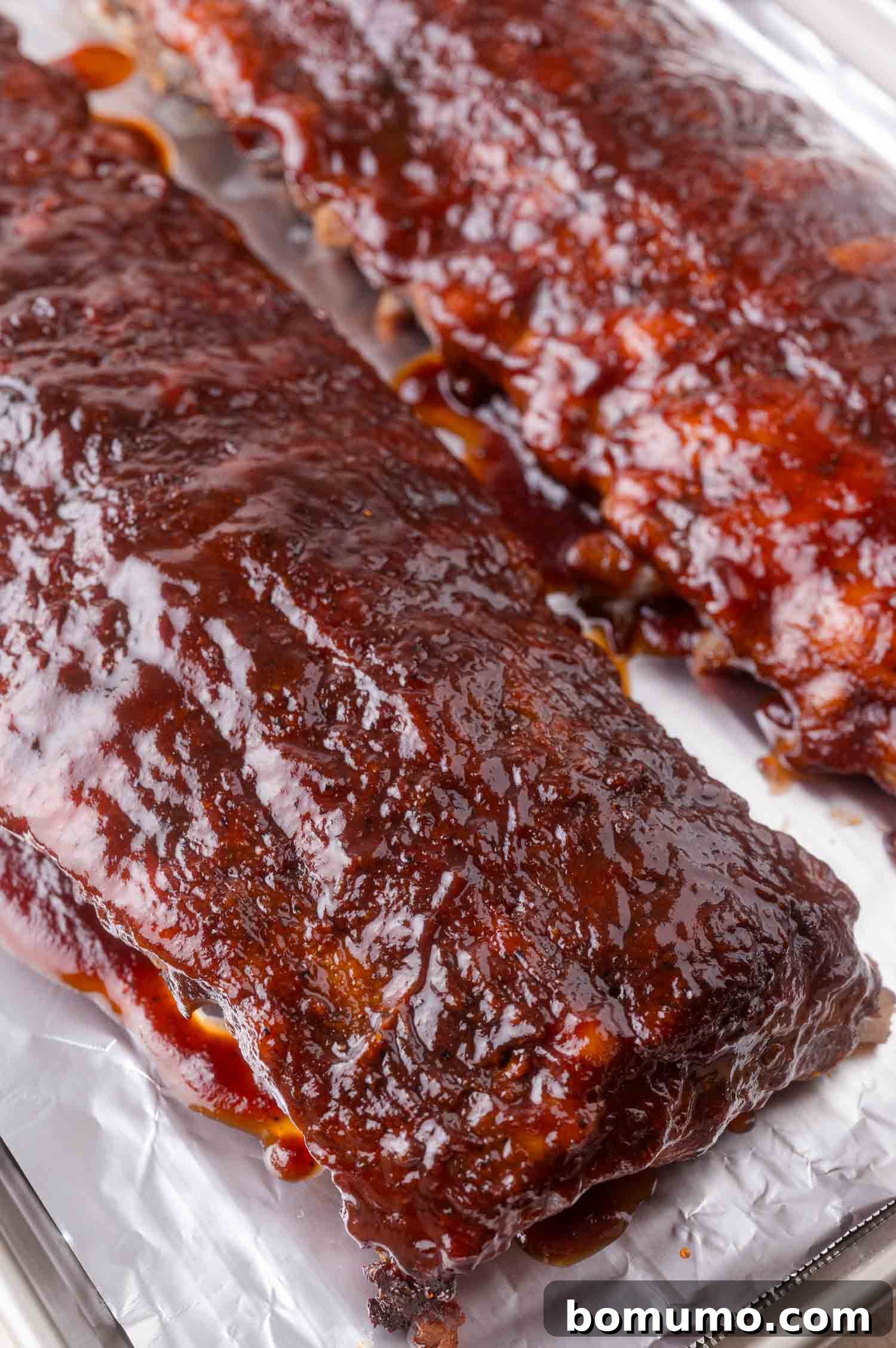 Two racks of barbecued ribs covered in glossy sauce rest on a foil-lined baking sheet.