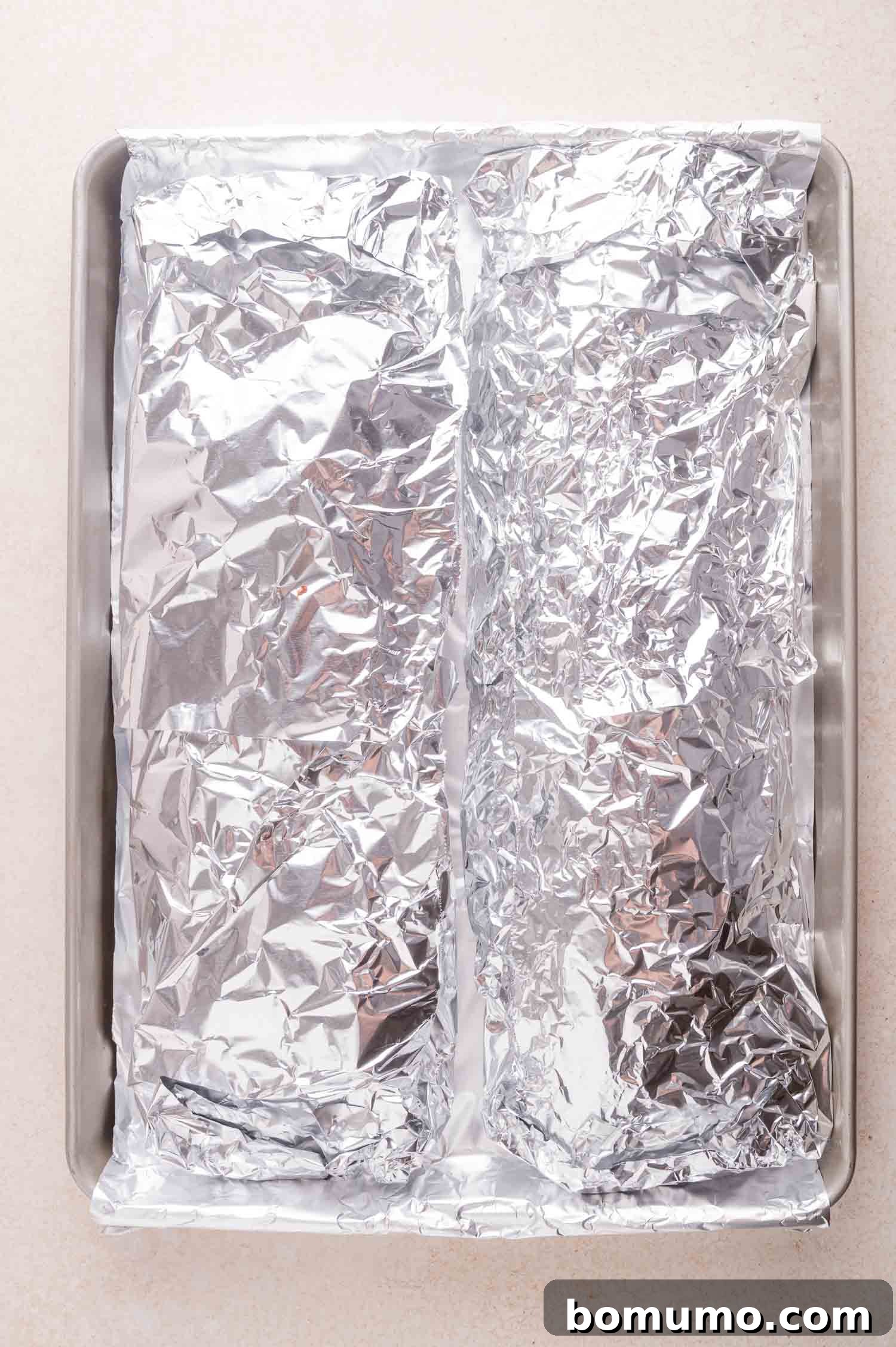 A baking sheet with two racks of ribs wrapped in aluminum foil, placed side by side on a light-colored countertop.