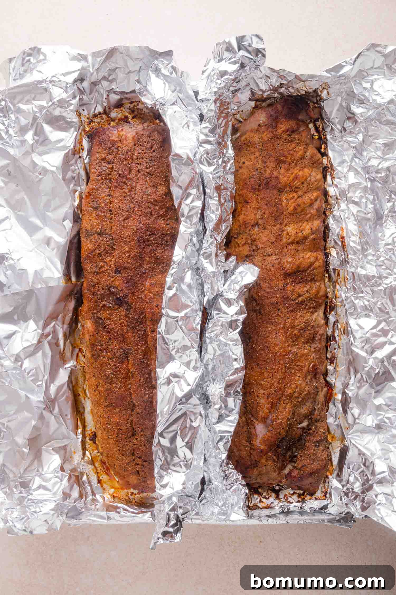 Two racks of seasoned, cooked ribs resting in aluminum foil on a light-colored surface.