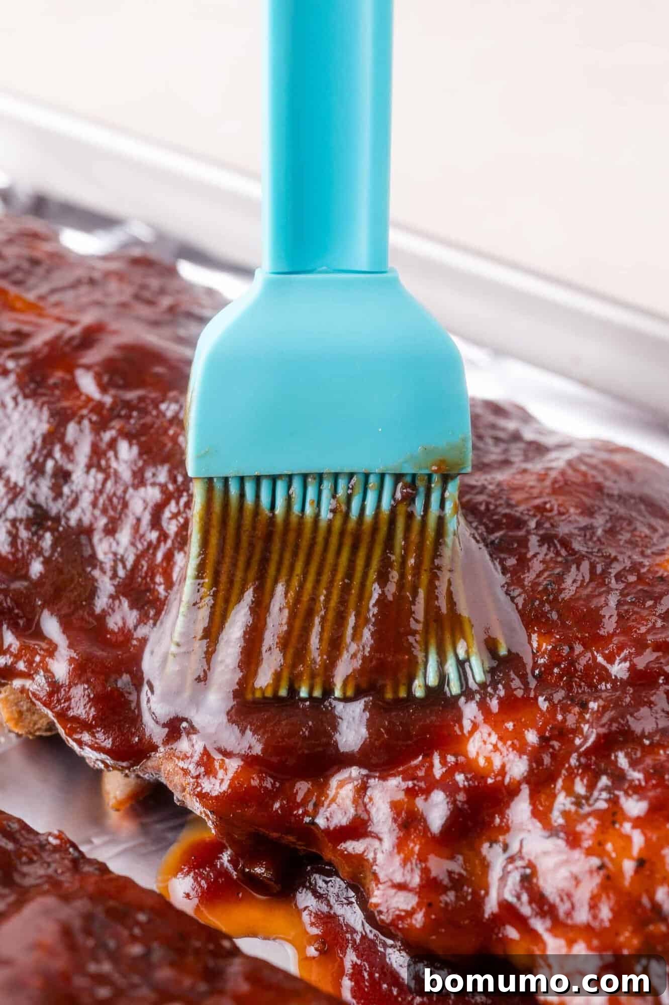 A close-up of a blue silicone brush applying barbecue sauce onto a rack of ribs on a metal tray.