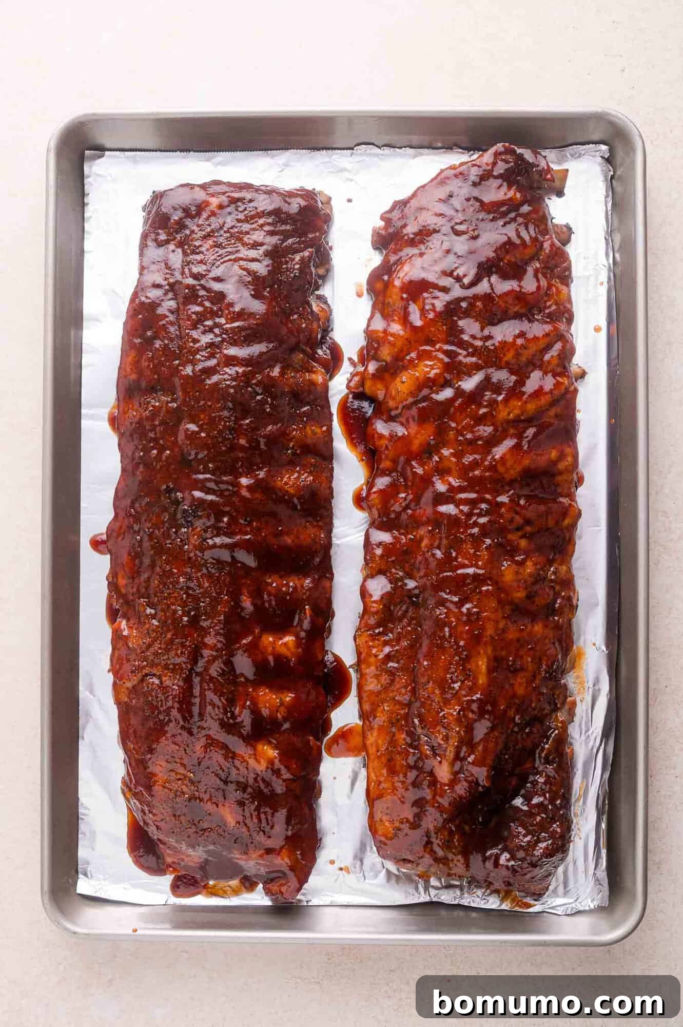 Two racks of barbecued ribs covered in sauce on a foil-lined baking tray.