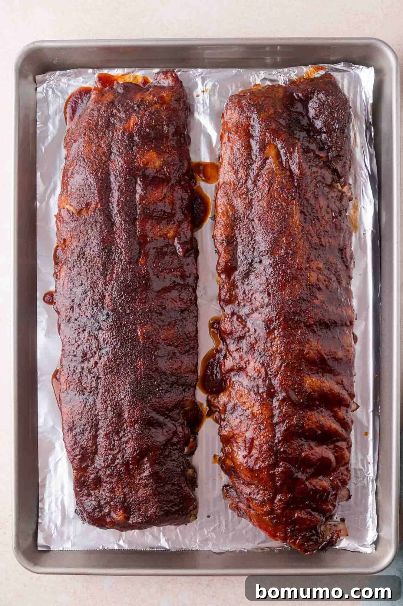 Two racks of cooked, glazed ribs rest on a foil-lined baking sheet.