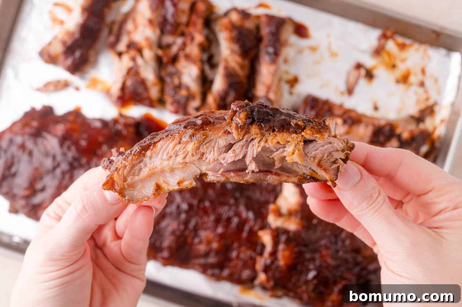 A person holds a single barbecued rib in front of a tray with more cooked ribs on parchment paper.
