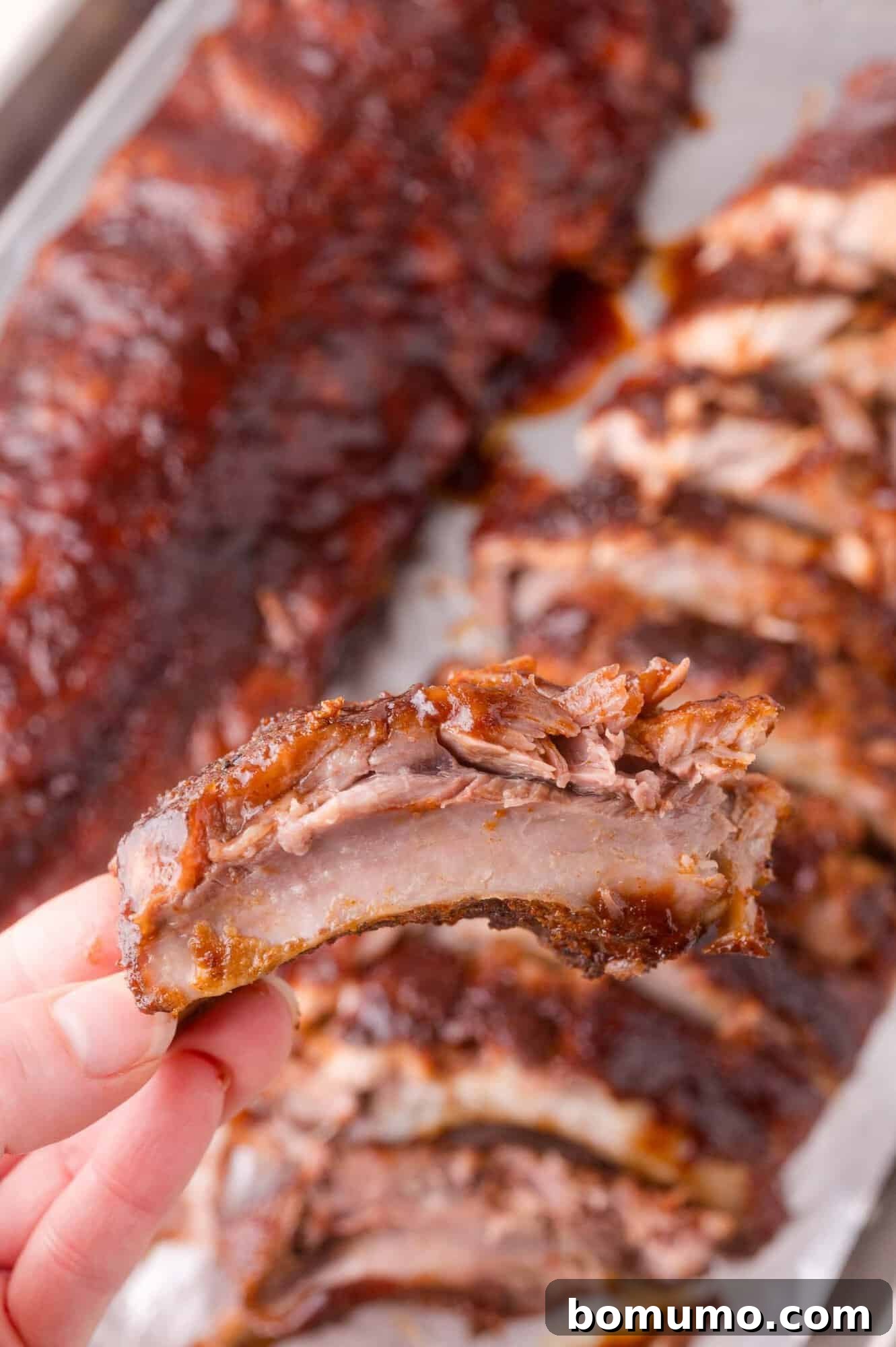 A hand holds a single, saucy barbecued rib with more sliced ribs and a whole rack in the background on a tray.