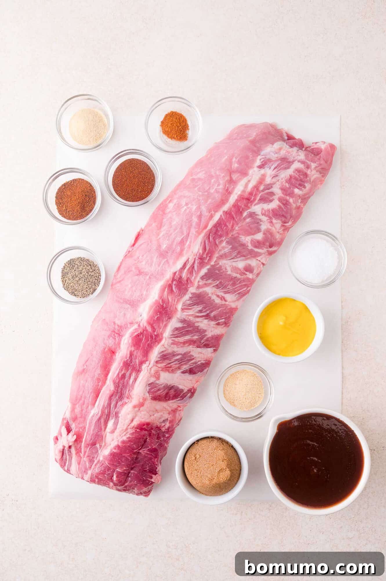 Uncooked rack of pork ribs on a white surface, surrounded by small bowls of spices, mustard, salt, pepper, sugar, and barbecue sauce.
