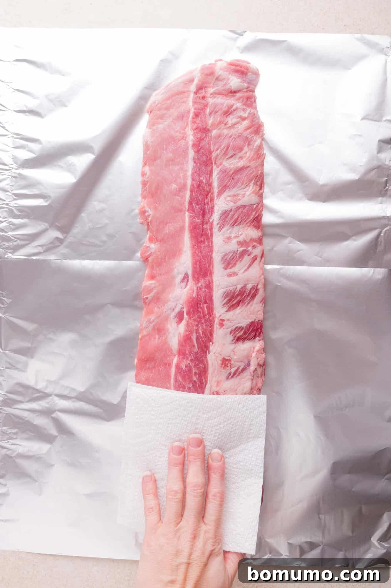 A hand using a paper towel to pat dry a raw rack of ribs placed on a sheet of aluminum foil.