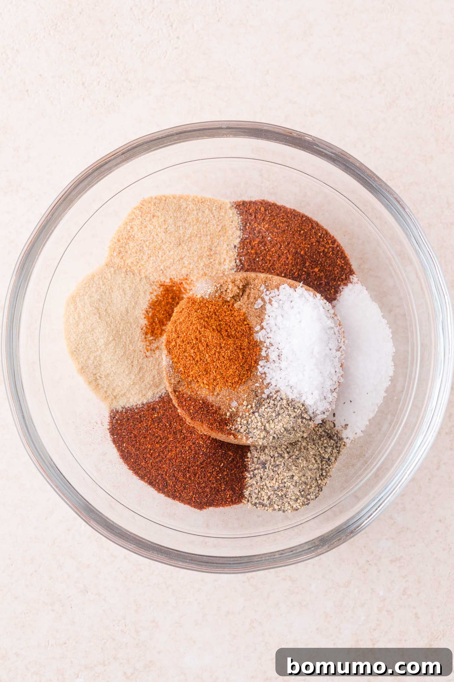A clear glass bowl containing separate piles of various spices, including salt, pepper, paprika, garlic powder, onion powder, and other seasonings, on a light surface.