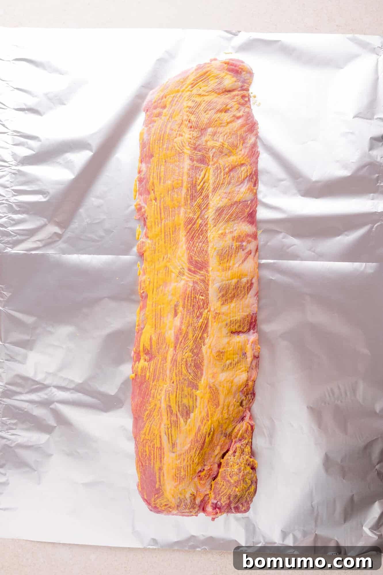 A rack of raw pork ribs coated with yellow mustard is placed on a sheet of aluminum foil.