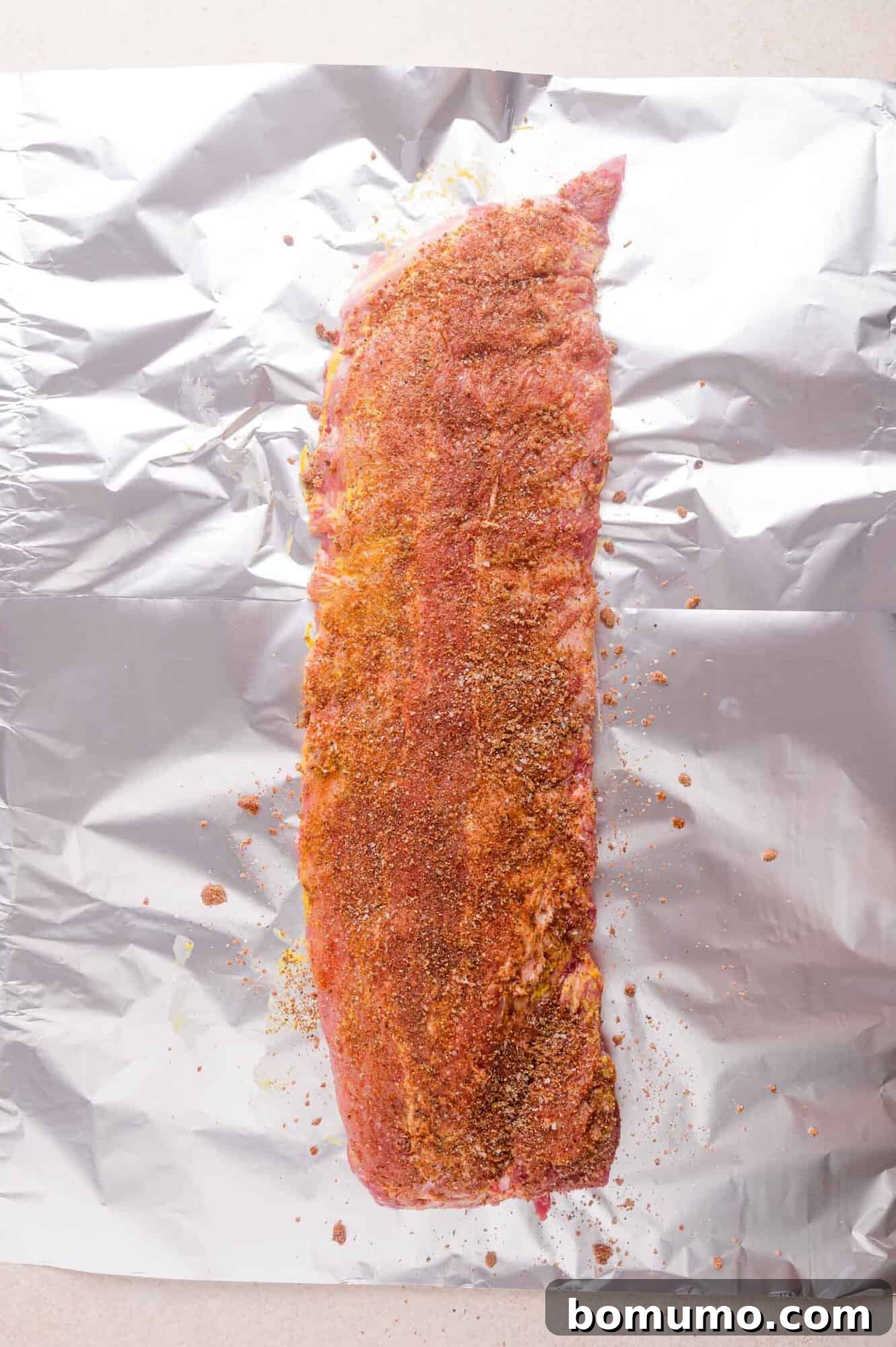 Raw pork ribs coated with dry seasoning sit on a sheet of aluminum foil, ready for cooking.