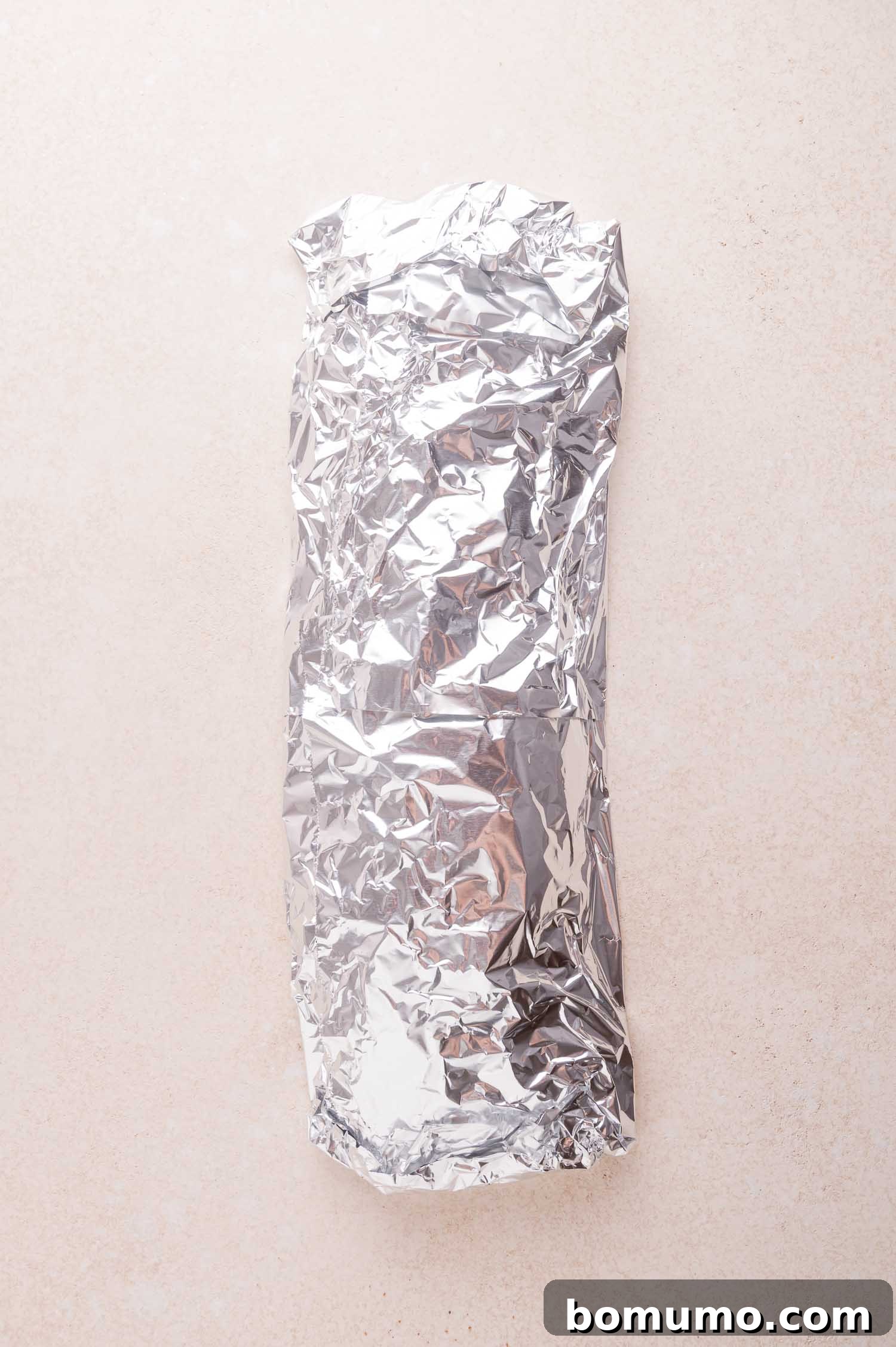 A rack of ribs wrapped tightly in crinkled aluminum foil, placed on a light-colored surface.