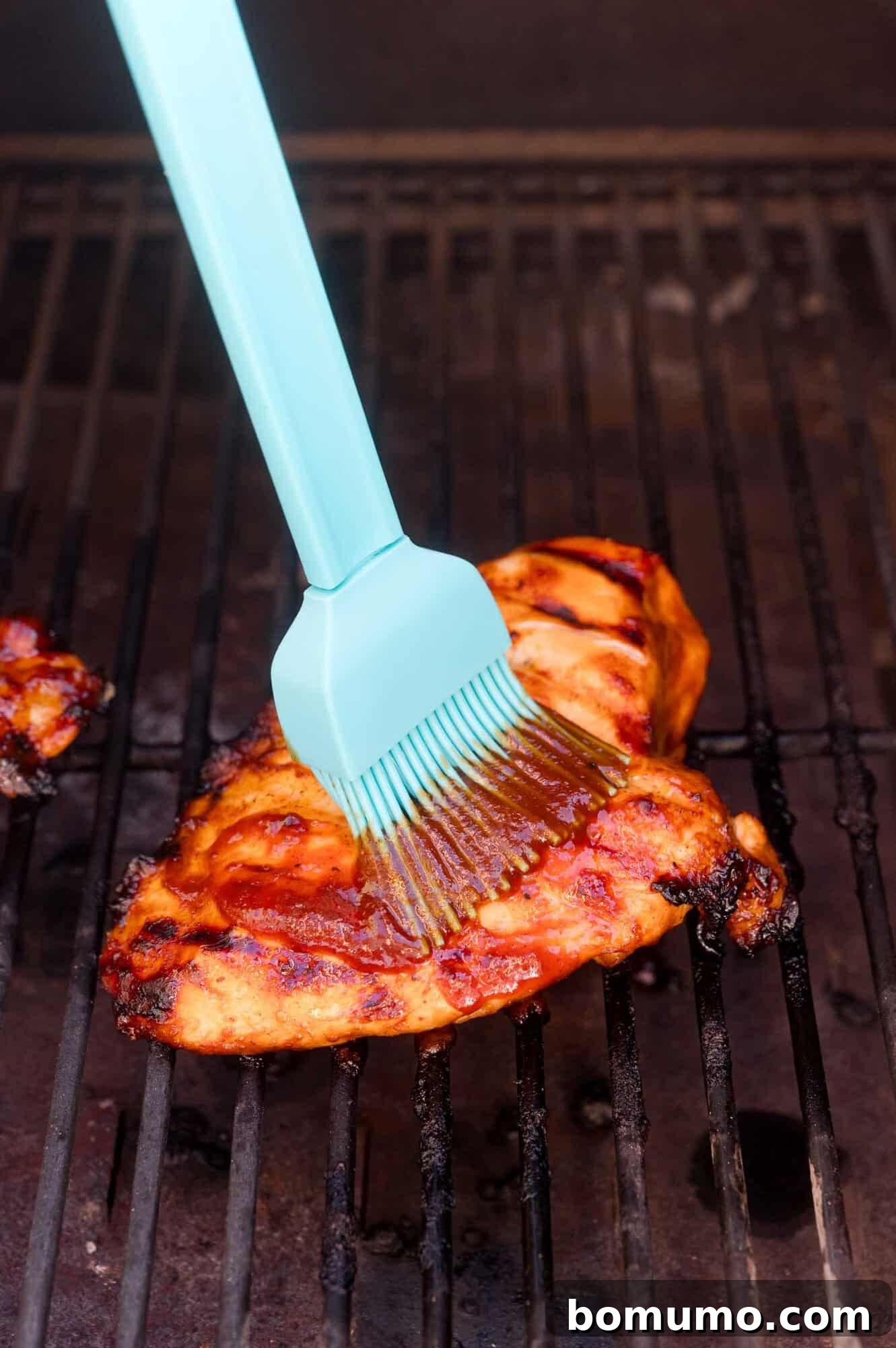 A blue silicone brush applies sauce to a grilled chicken breast on a barbecue grill.