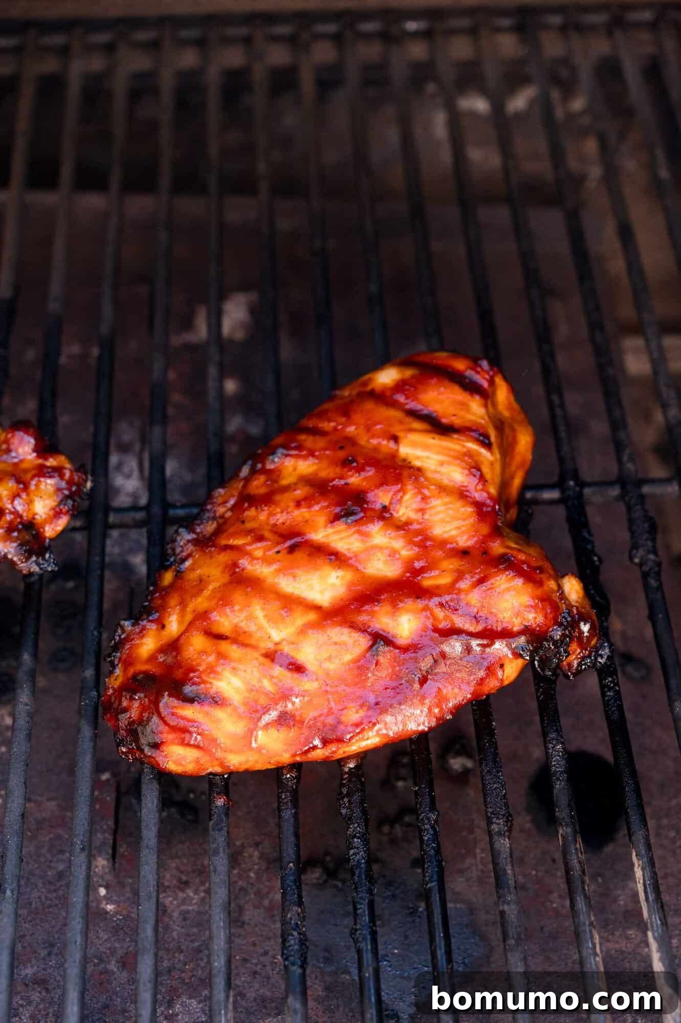 A grilled chicken breast coated in barbecue sauce sits on a hot grill with visible grill marks.