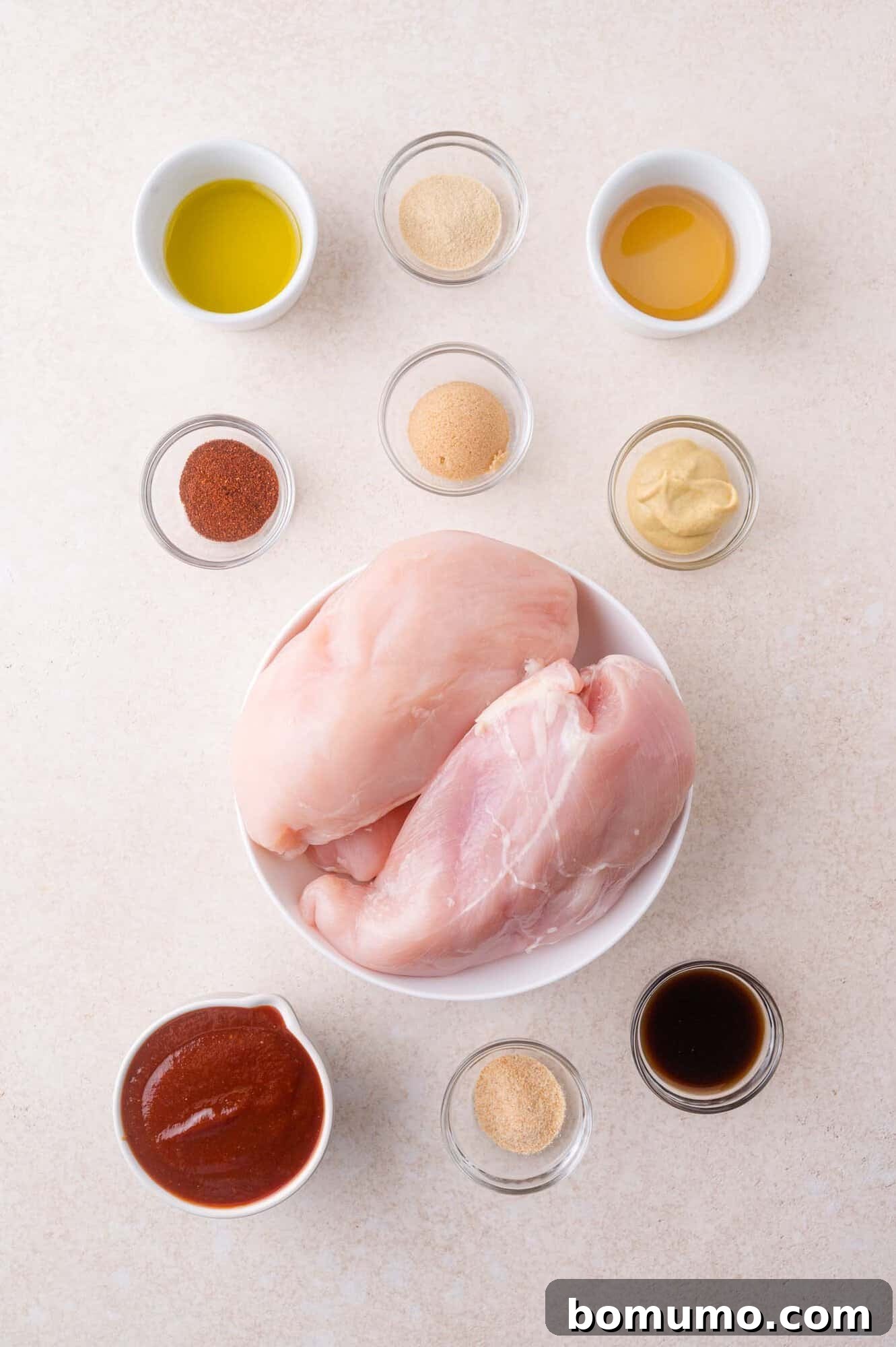 Raw chicken breasts in a bowl surrounded by small bowls containing various spices, sauces, and seasonings arranged on a light surface.