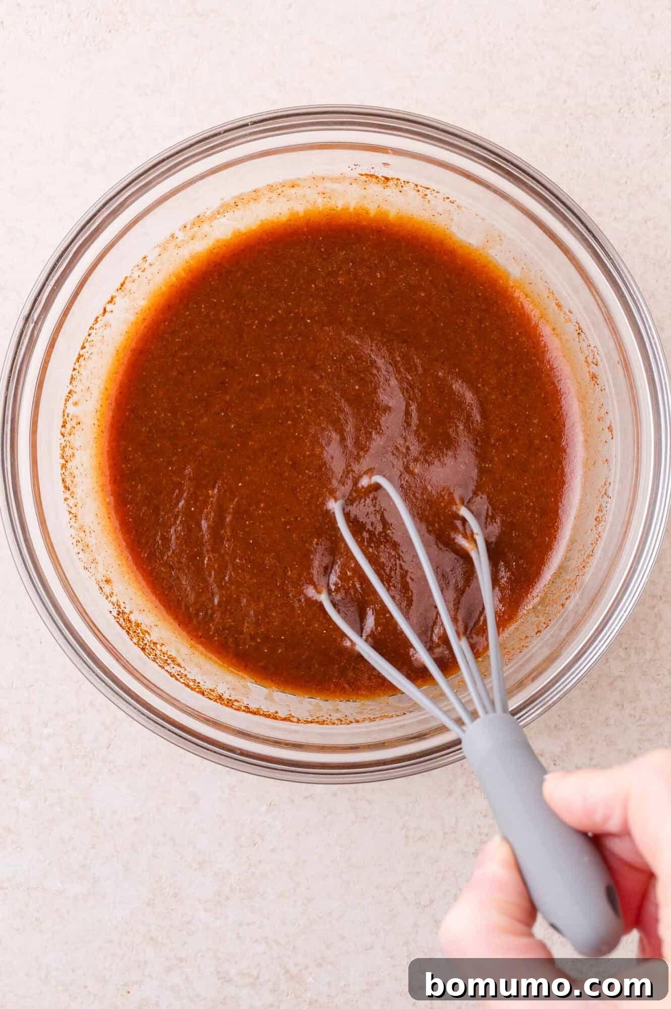 A hand holding a whisk mixes barbecue marinade in a clear glass bowl on a light countertop.