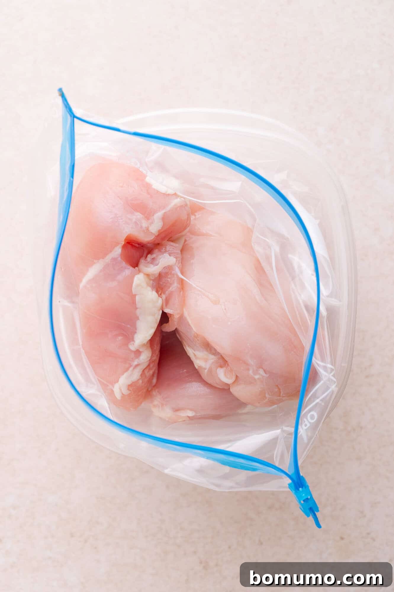 Raw chicken breasts in a clear resealable plastic bag on a light-colored surface.