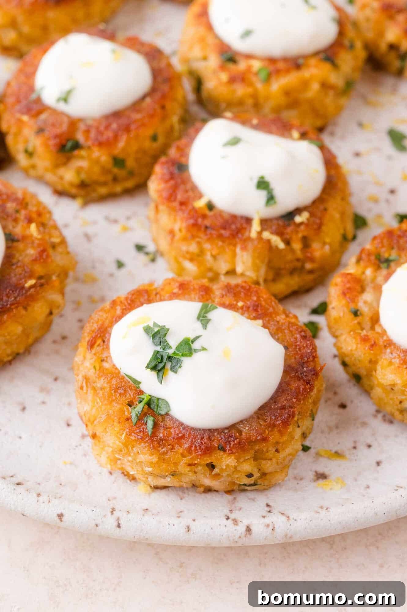 Several golden-brown crab cakes topped with dollops of lemon aioli and sprinkled with chopped herbs are arranged on a speckled white plate.