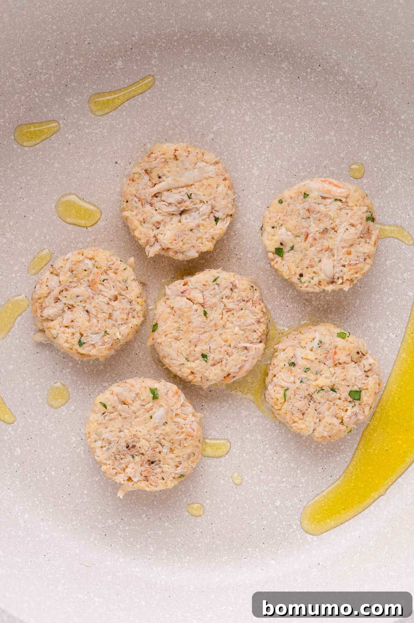 Six uncooked crab cakes arranged in a frying pan with a drizzle of oil.
