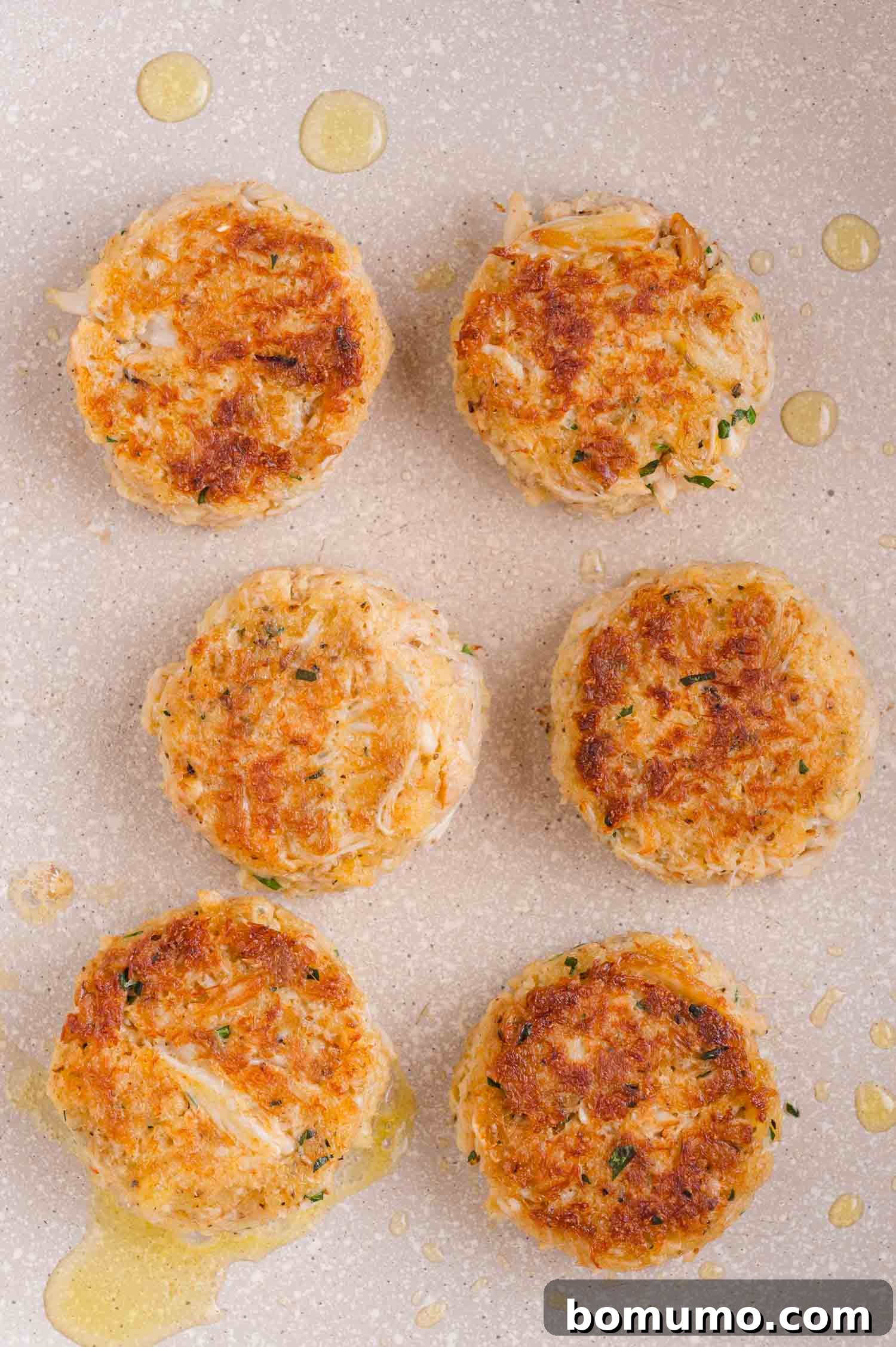 Six golden-brown crab cakes are arranged in a skillet with small drops of oil around them.