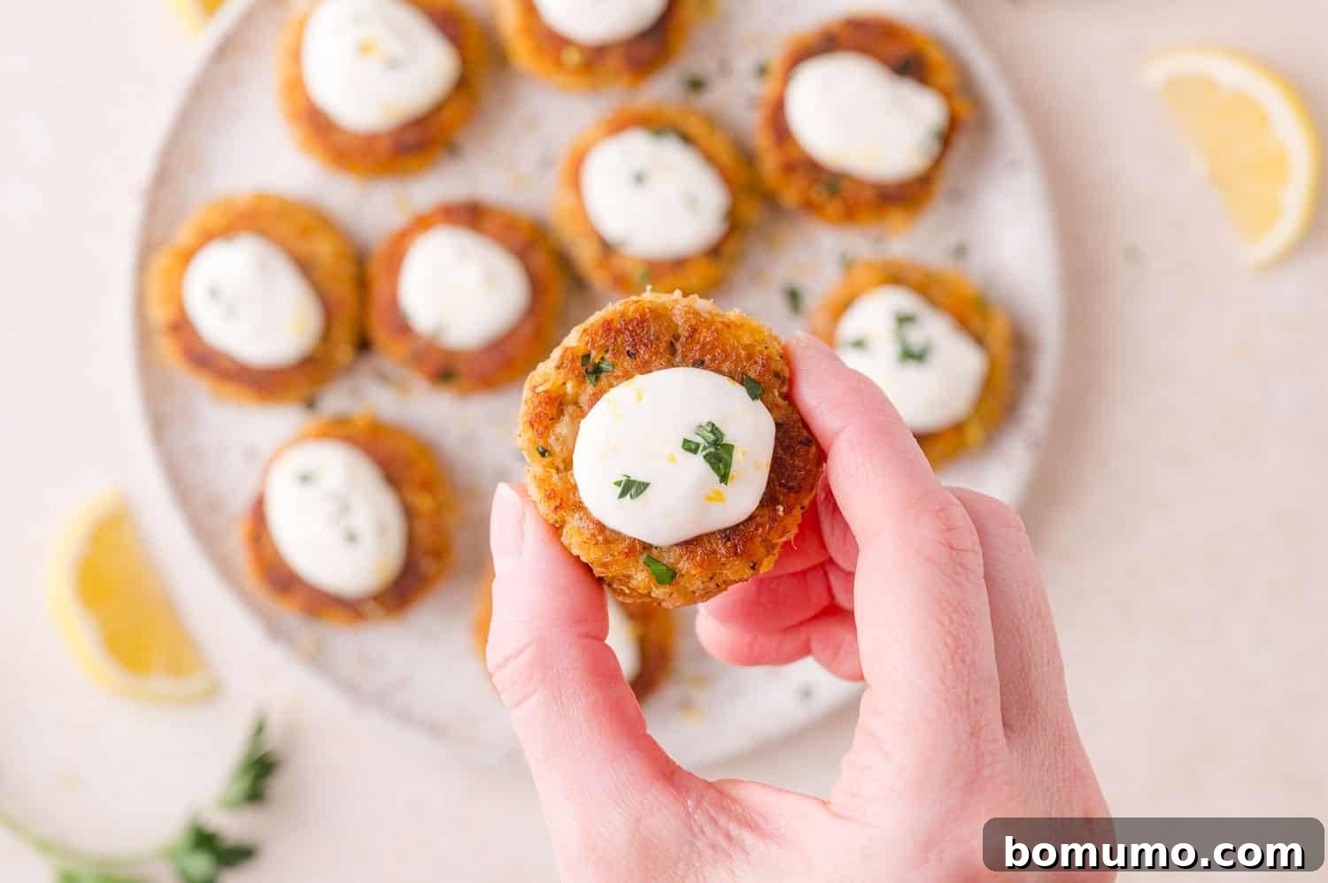 A hand holding a small, round crab cake topped with aioli and herbs, with more appetizers on a plate in the background. Lemon wedges and herbs are scattered nearby.