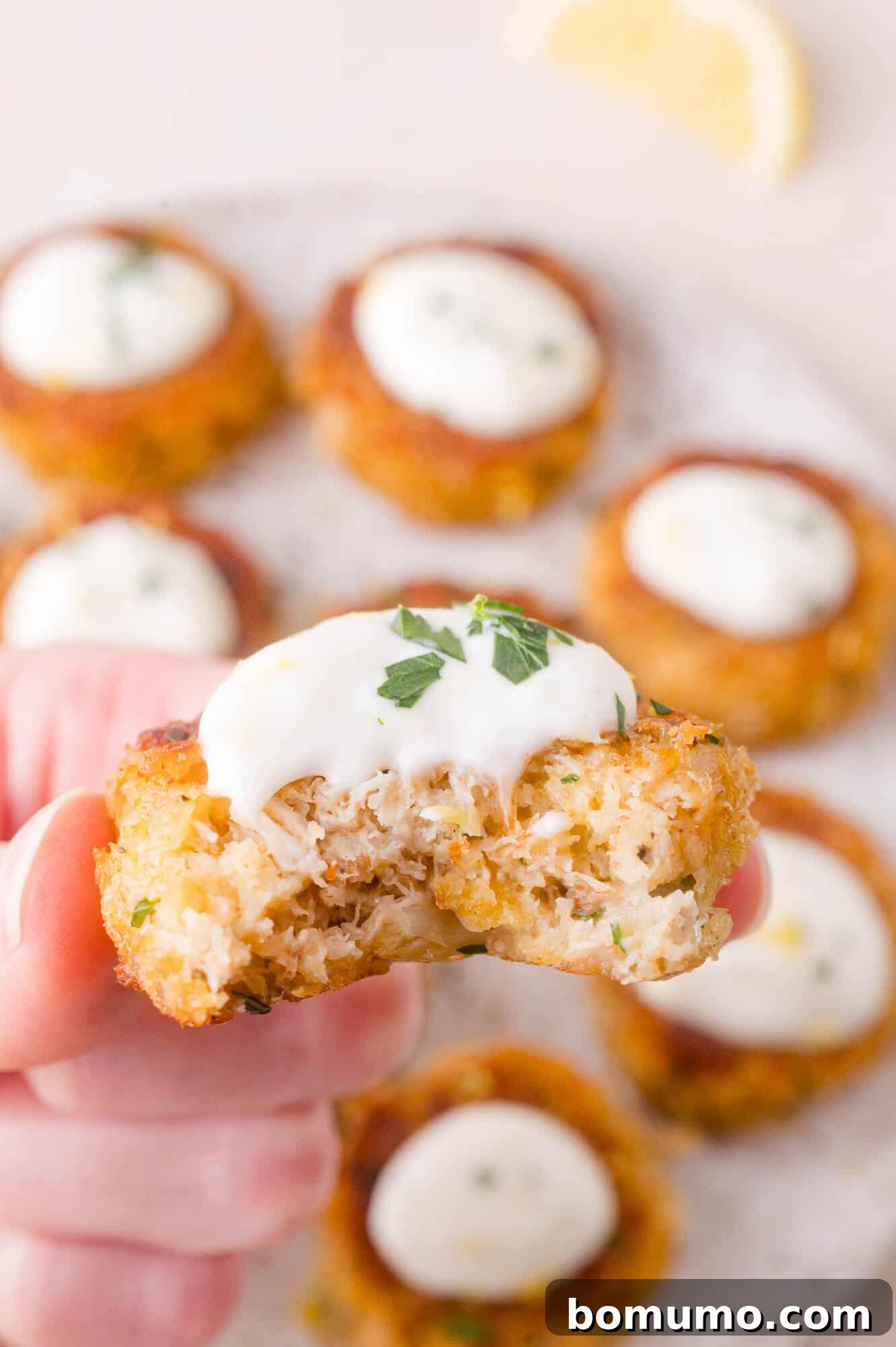 A hand holds a partially eaten mini crab cake topped with white aioli; several whole crab cakes with sauce are visible in the background.