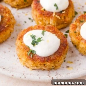 Close-up of golden brown crab cakes topped with a dollop of white aioli and garnished with chopped fresh herbs on a speckled plate.
