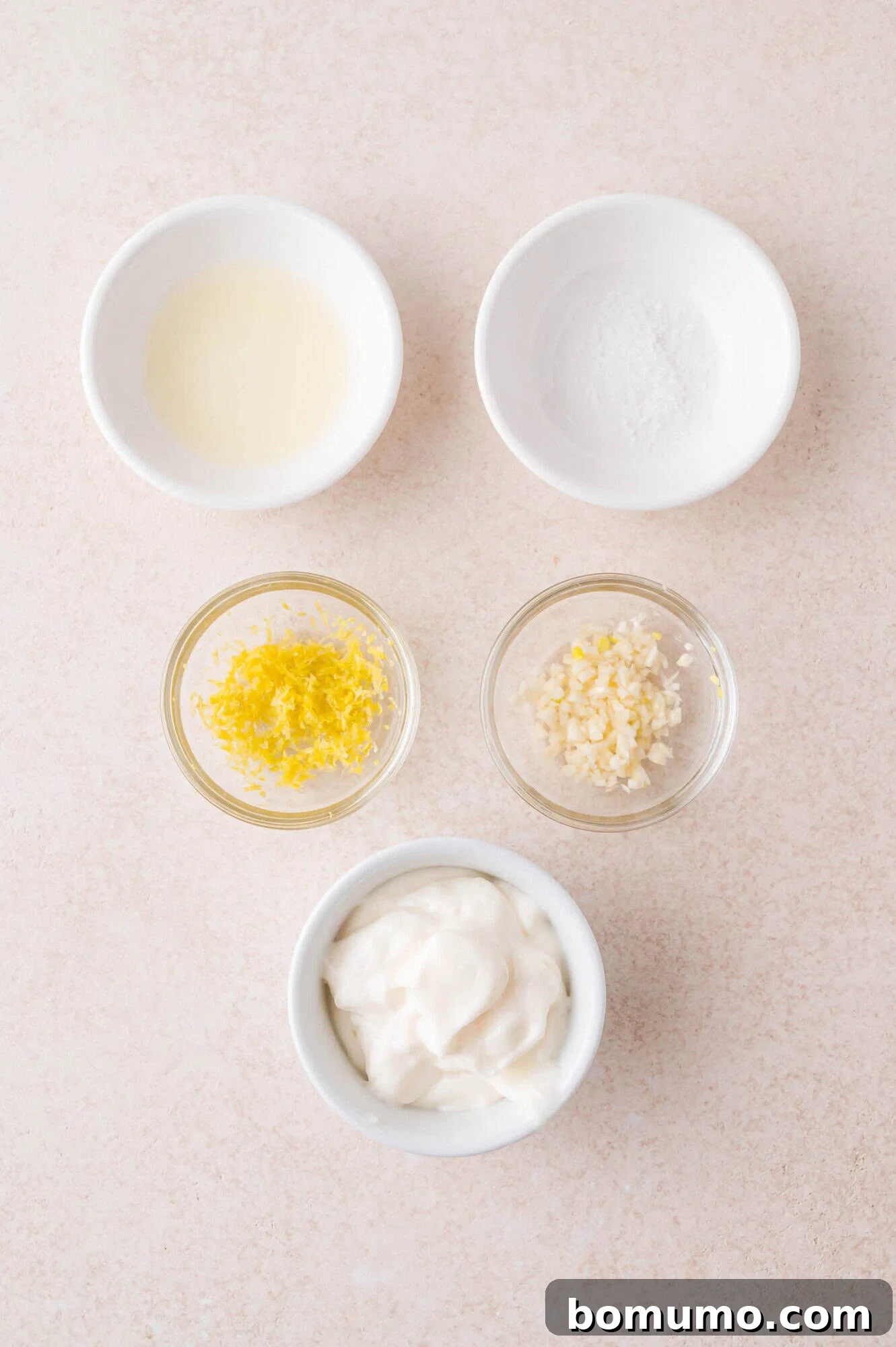 Five small bowls containing lemon juice, salt, lemon zest, minced garlic, and mayonnaise arranged on a light surface.