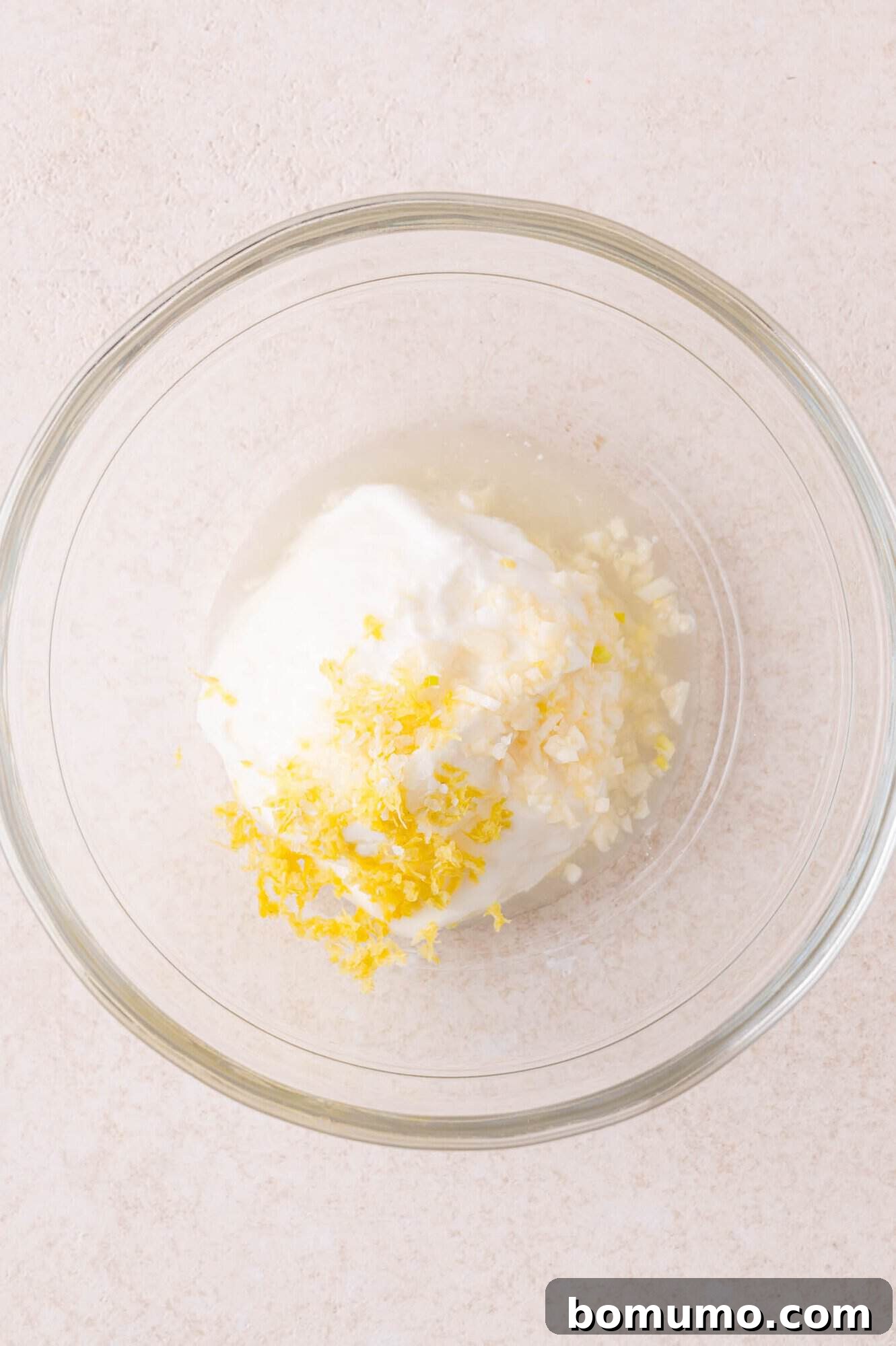 A glass bowl containing yogurt, minced garlic, lemon zest, and lemon juice on a light-colored surface.