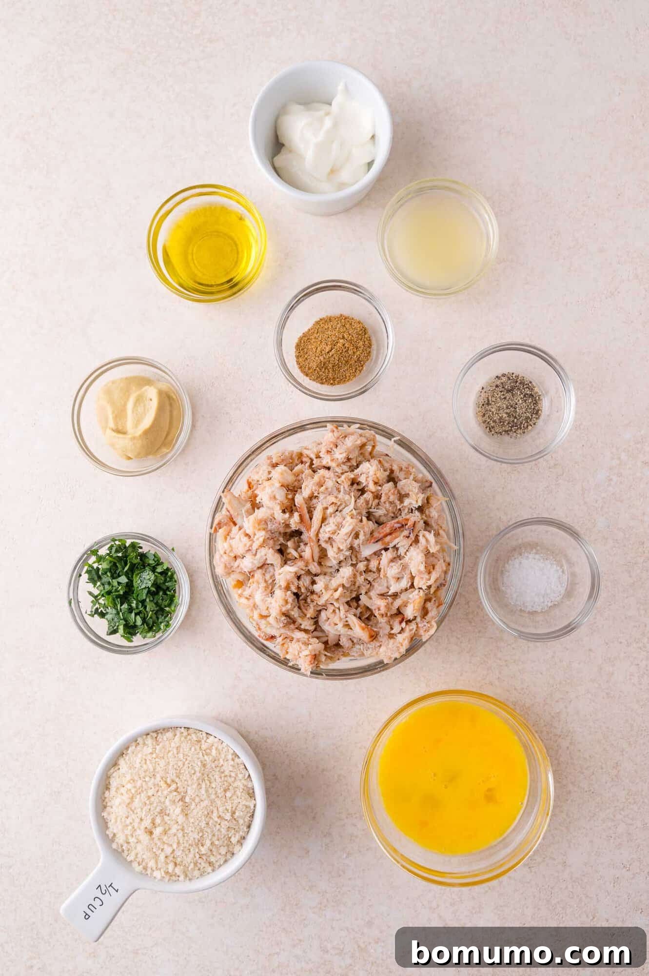 Overhead view of separate bowls containing crab meat, panko breadcrumbs, egg, mayonnaise, mustard, parsley, oil, lemon juice, and seasonings arranged on a light surface.
