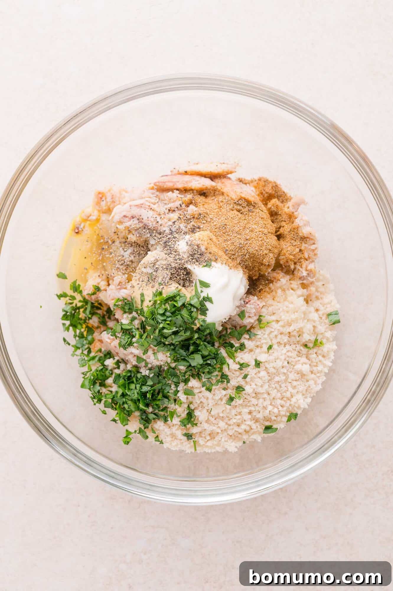 A glass bowl containing crab meat, breadcrumbs, chopped herbs, seasoning, an egg, and a dollop of mayonnaise on a light surface.