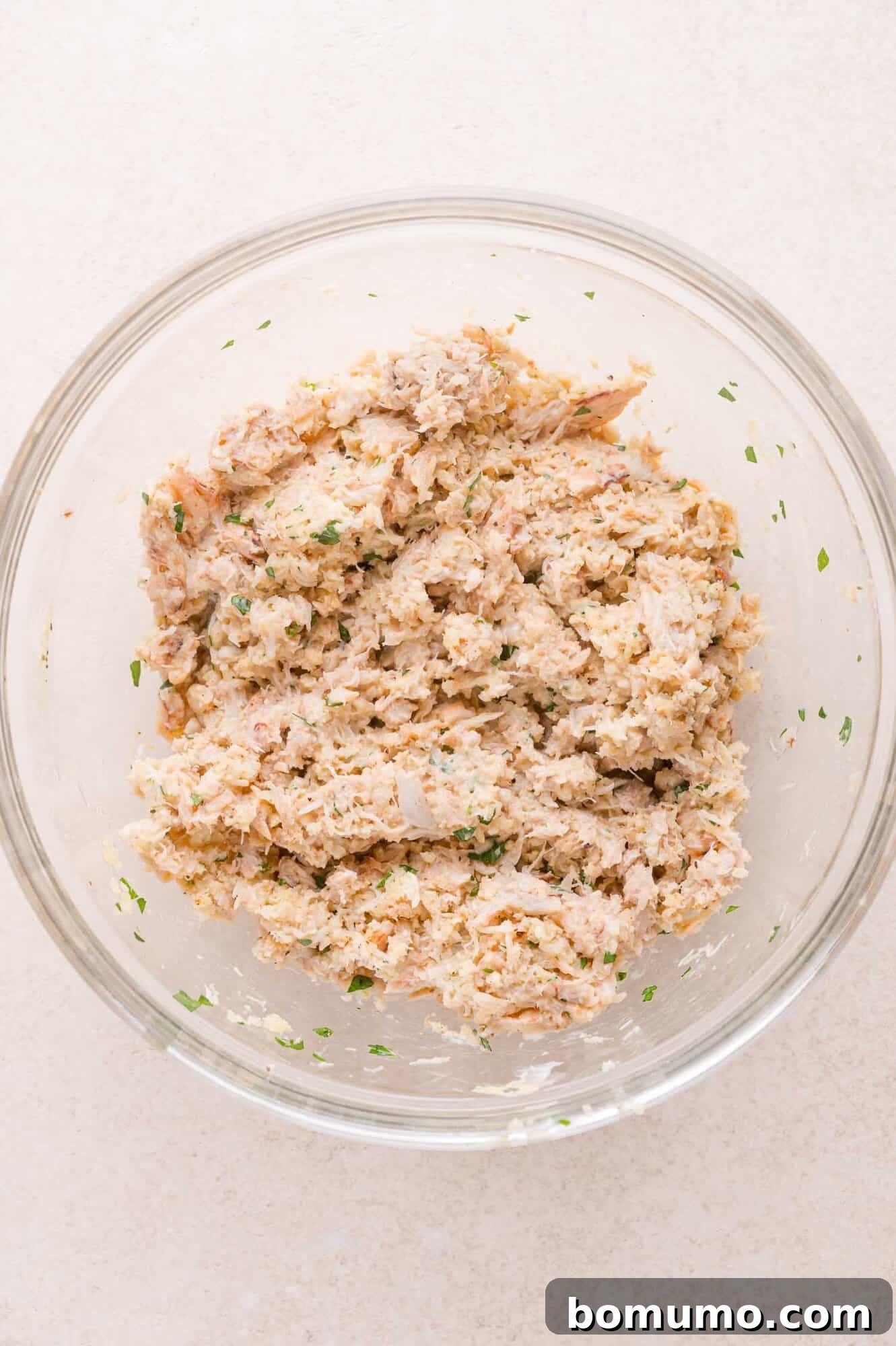 A clear glass bowl containing a mixture of crab, herbs, and other finely chopped ingredients on a light surface.