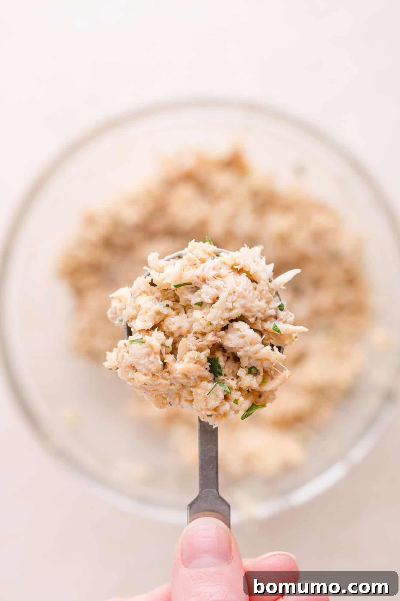 A hand holds a spoonful of a mixed crab filling above a glass bowl containing more of the mixture.