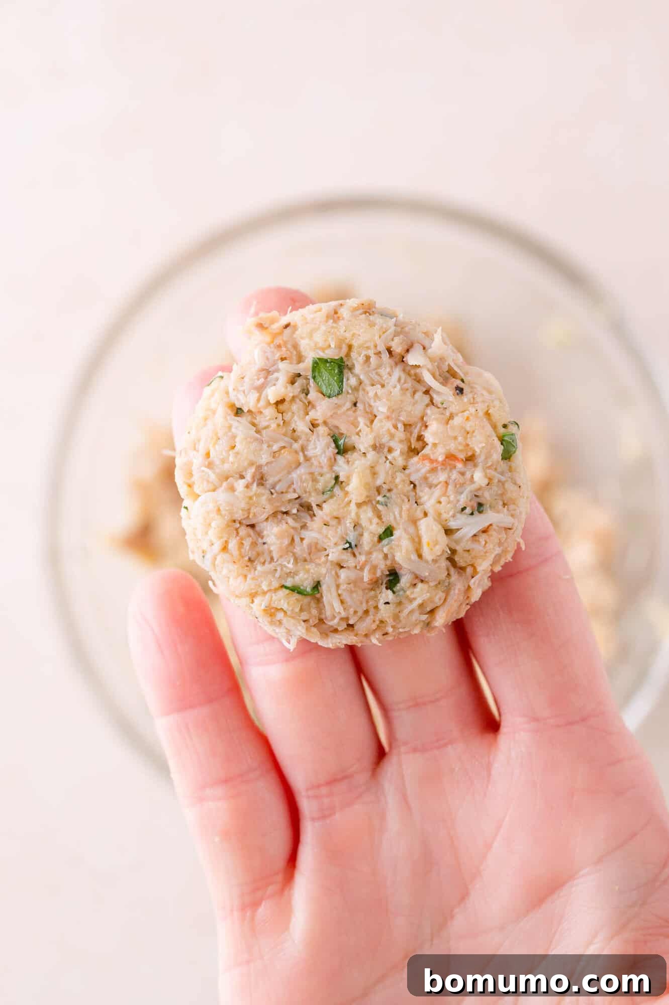 A hand holds an uncooked crab cake patty above a glass bowl containing the crab mixture.