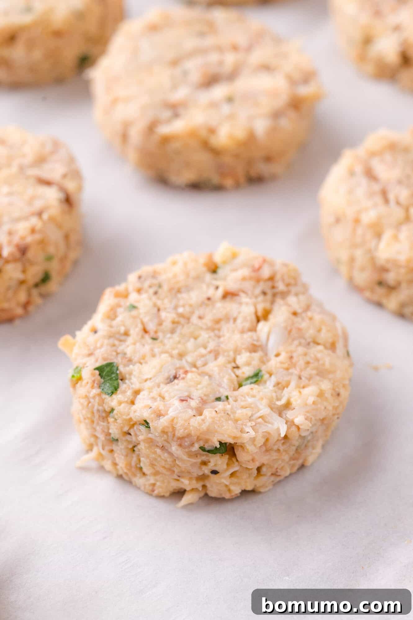 Uncooked crab cakes formed into round patties rest on a sheet of parchment paper.