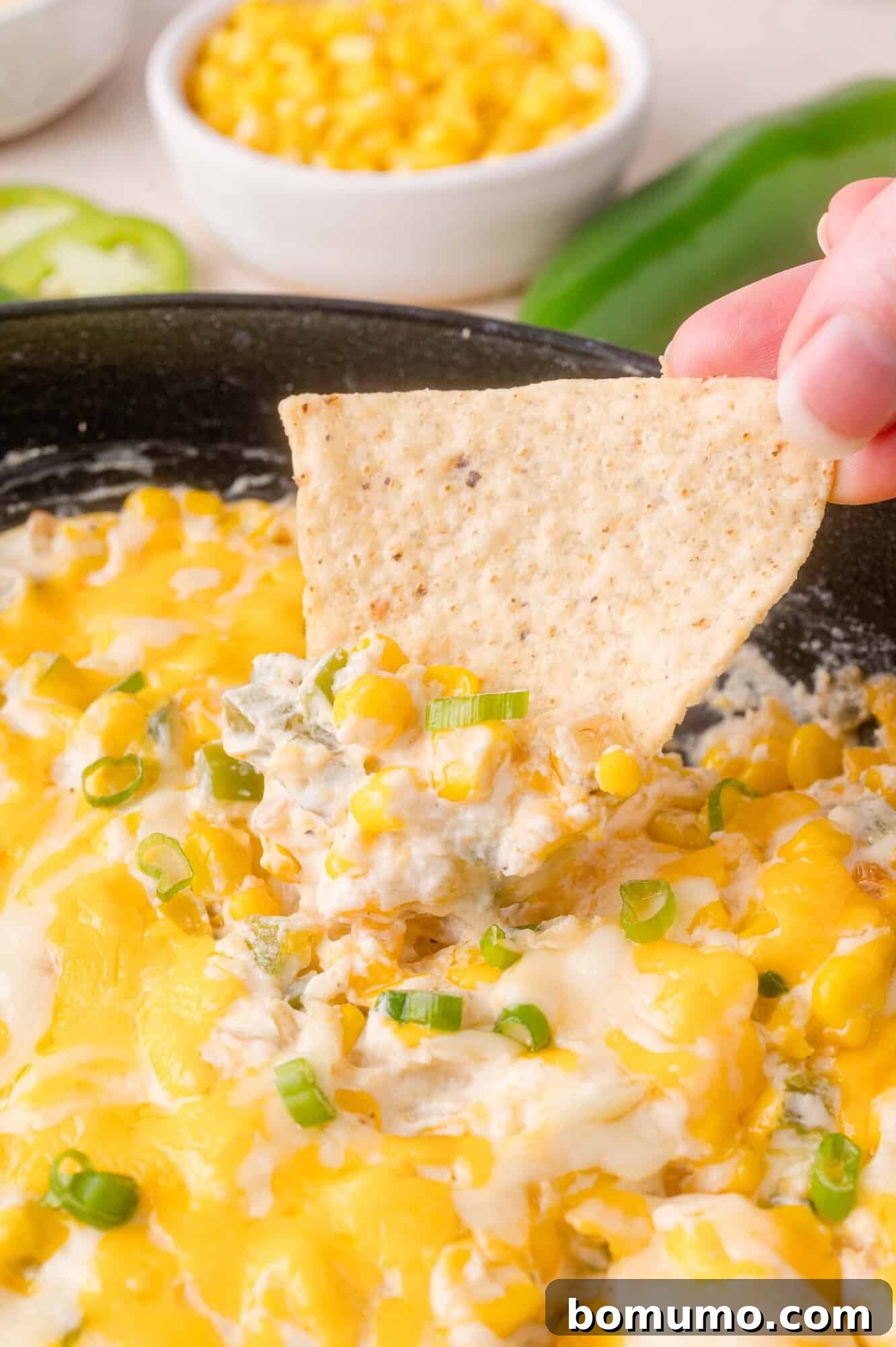 A hand holds a tortilla chip dipped into a cheesy corn and jalapeño dip, topped with green onions, with a bowl of corn in the background.