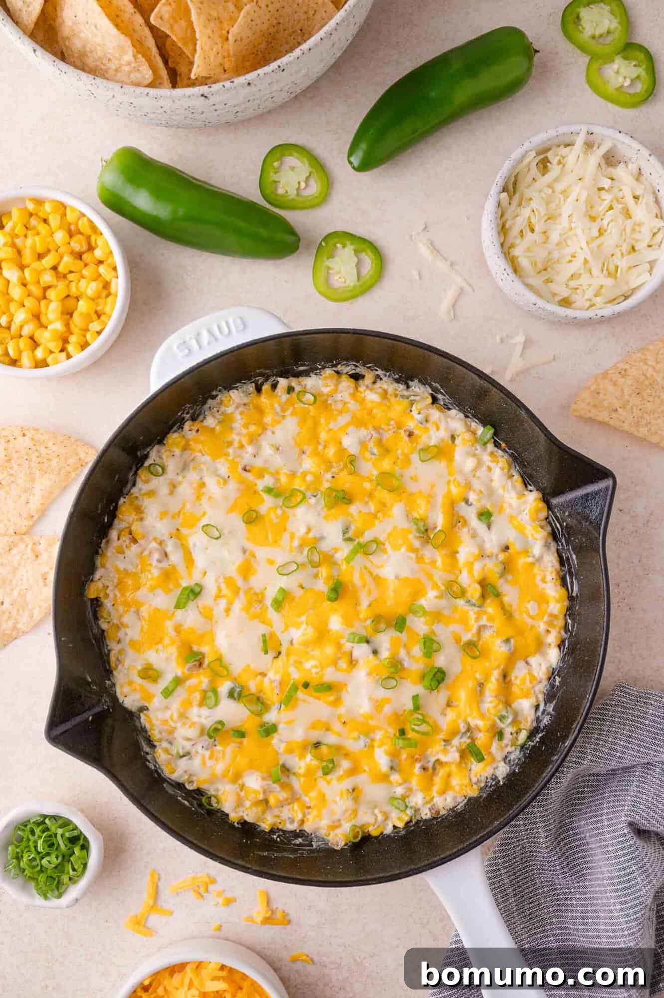 A skillet of melted cheese dip garnished with chopped green onions, surrounded by tortilla chips, corn, shredded cheese, and sliced jalapeños.