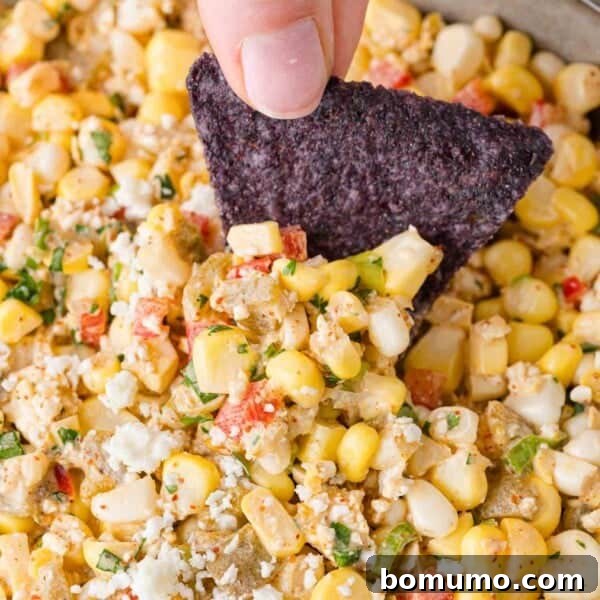 A blue tortilla chip is dipped into a mixture of corn, diced red and green onions, and crumbled cheese.