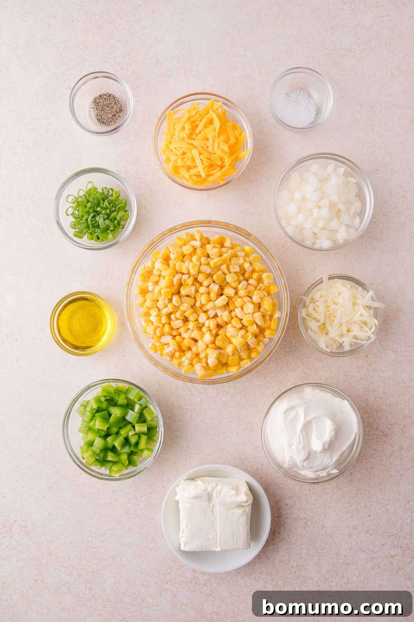Overhead view of bowls containing corn, shredded cheese, diced onion, chopped jalapeno pepper, green onions, cream cheese, sour cream, oil, salt, and pepper on a light surface.