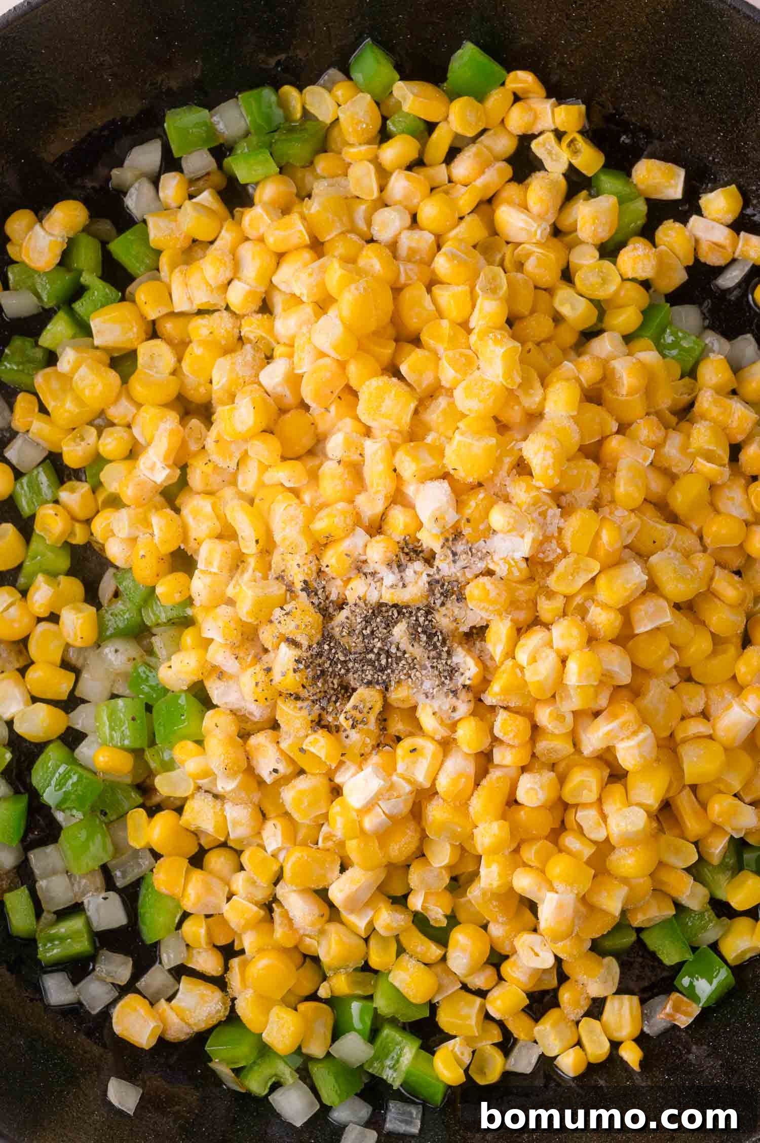 Corn, diced pepper, and chopped onion cooking in a skillet, topped with ground black pepper and seasoning.