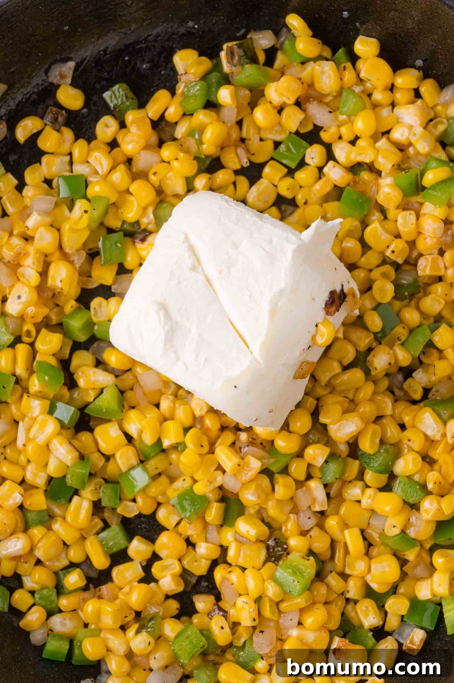 Corn kernels and chopped jalapenos in a pan with a block of cream cheese on top, ready to be mixed.